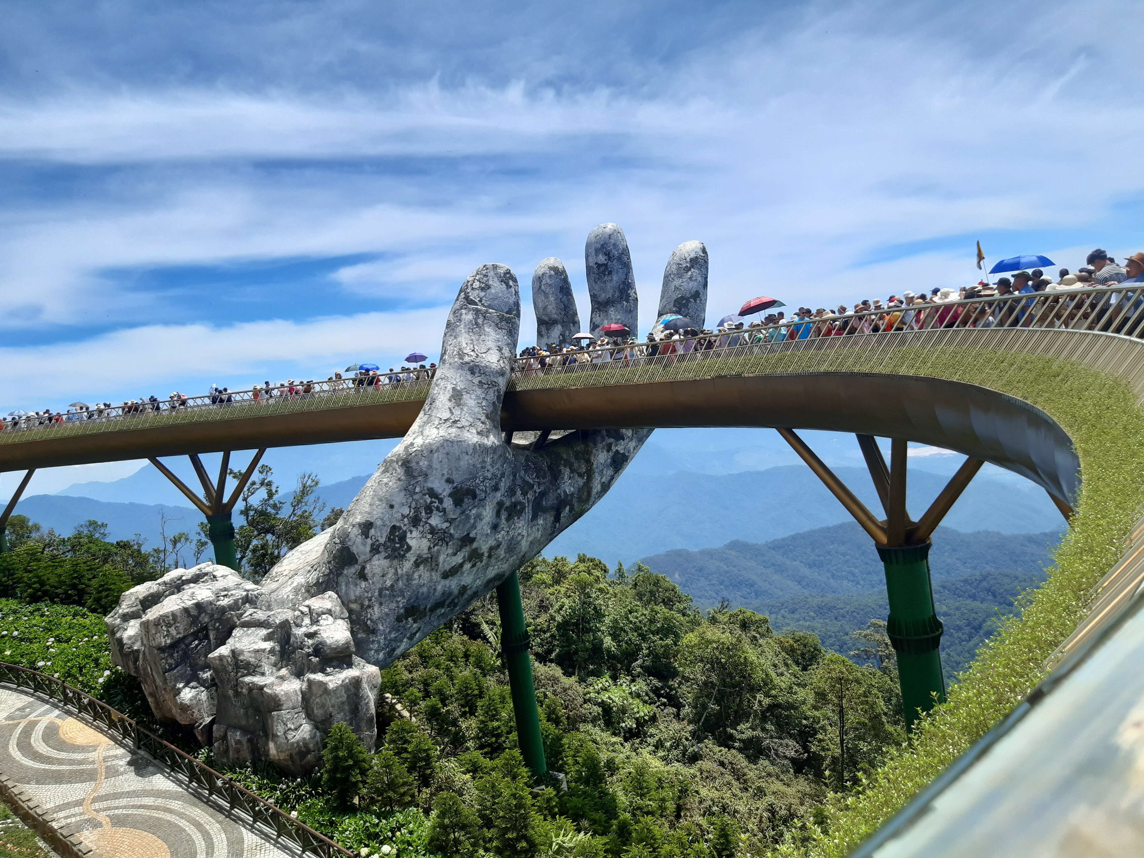 The Golden Bridge in Da Nang, Vietnam, supported by two giant stone hands, with crowds of visitors walking along the curved pedestrian walkway against a backdrop of lush mountains and blue sky.